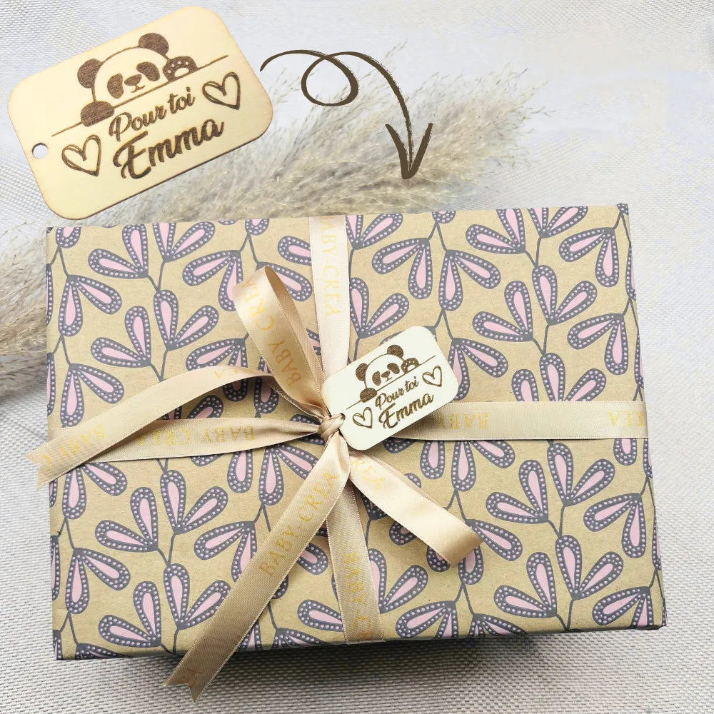 Cadeau de naissance personnalisé pour bébé fille, emballé avec soin dans un papier fleuri beige et rose, ruban satiné BABY-CREA et étiquette gravée au prénom "Emma". Idéal pour un coffret naissance élégant, prêt à offrir dès la maternité.